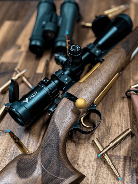 Lunetă Falke pe o carabină de vânătoare cu pat din lemn, cartușe puse pe masă. Foto: Rigad