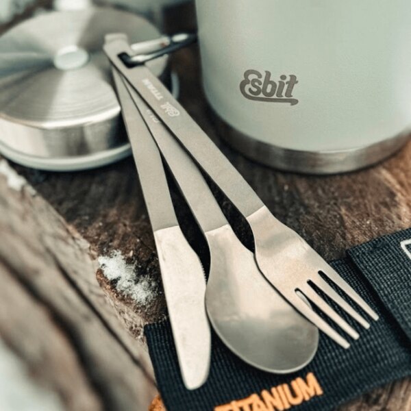 Furculiță, cuțit și lingură din titan Esbit lângă un termos de exterior pe un suport de lemn. Foto: Rigad