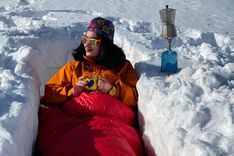 Femeia cu o cană de tablă stă într-un șanț acoperită cu un sac de dormit de iarnă. Sursa: https://www.pexels.com/cs-cz/foto/ryma-studeny-snih-zena-10284511/