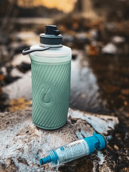 Sticla pliabilă Flux™ HydraPak® cu filtru de apă montat HydraPak® (28 mm) așezată pe o piatră lângă un pârâu. Foto: Rigad