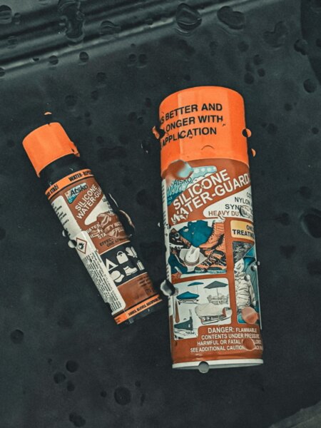Spray-uri impermeabile Atsko Silicone Water-Guard așezate pe o țesătură închisă la culoare cu picături de apă.