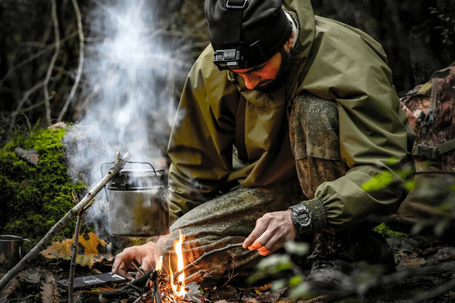 Bărbat într-o jachetă din membrană lângă foc în pădure. Sursa: https://www.pexels.com/photo/kneeling-man-starting-fire-in-a-fireplace-15538110/