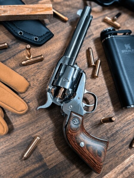 Un revolver cu acțiune simplă Ruger cu țeavă lungă și mâner de lemn așezat între gloanțe și echipament de exterior.