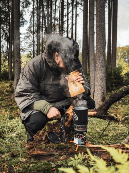 Un bărbat într-o geacă groasă stă în genunchi în pădure lângă un aragaz Jetboil, din care se ridică aburi. Pregătește o masă caldă sau o băutură ca sursă de energie și căldură în timpul unei excursii de toamnă. Foto: Rigad