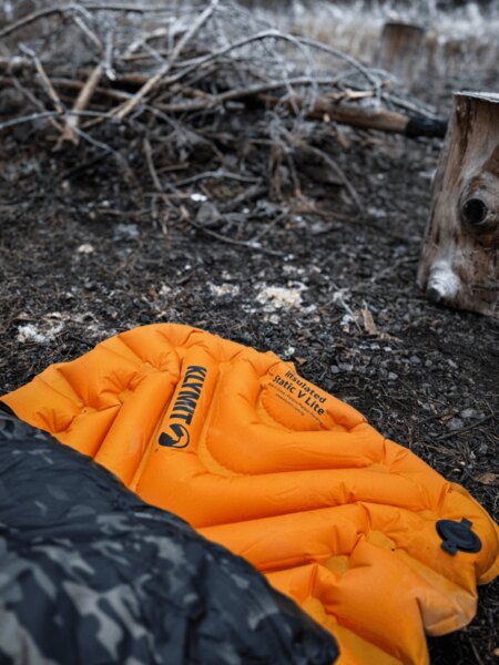 Saltea gonflabilă Klymit Insulated Static V Lite de culoare portocalie pe pământ lângă foc, parțial acoperită de un sac de dormit cu model de camuflaj. Izolație împotriva frigului și umezelii la dormit în teren. Foto: Rigad