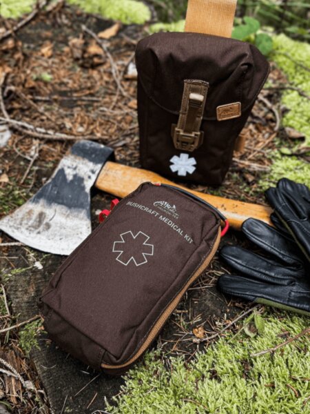 Kit medical Helikon-Tex Bushcraft în husă maro, așezat în pădure lângă un topor și mănuși – kit medical pentru bushcraft și activități în aer liber. Foto: Rigad.