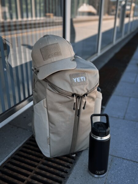 Rucsac verde YETI Crossroads 27 l cu șapcă, ochelari de soare și recipient negru YETI Rambler.