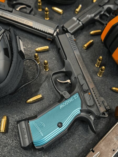 Pistolă sportivă CZ Shadow 2 cu mânere din dural albastru între cartușe și echipamente de tir.